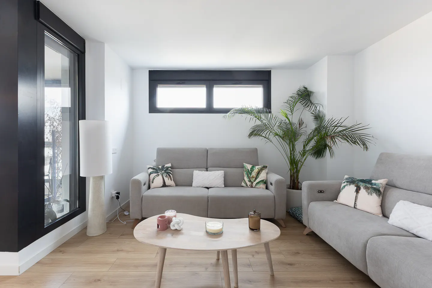Bright living room with light wood floors, gray sofas with palm tree pillows, and a light wood coffee table. A large potted palm sits near a window.