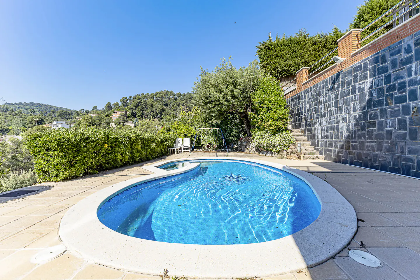 Kidney-shaped pool with blue water, surrounded by stone patio, green foliage, and a stone wall under a clear blue sky.