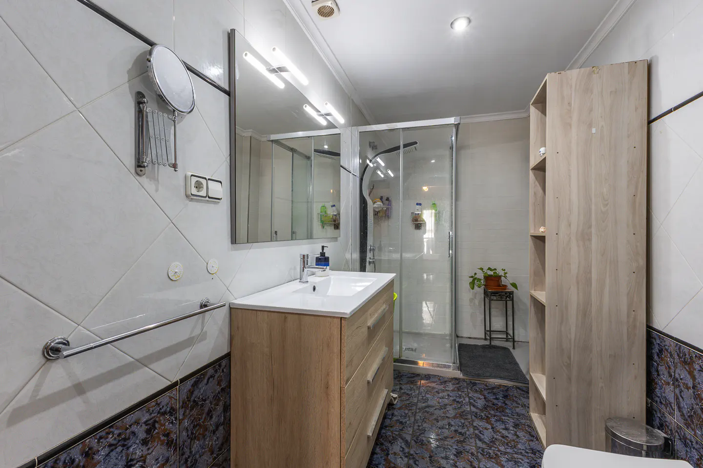 Bright bathroom with a wood vanity, white sink, and glass shower. Blue and white patterned tile floor.