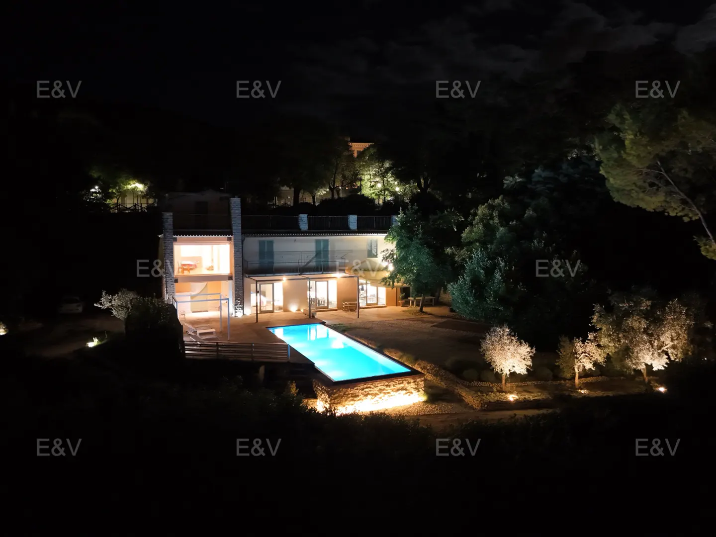 A lit two-story house with a blue pool at night, surrounded by trees.