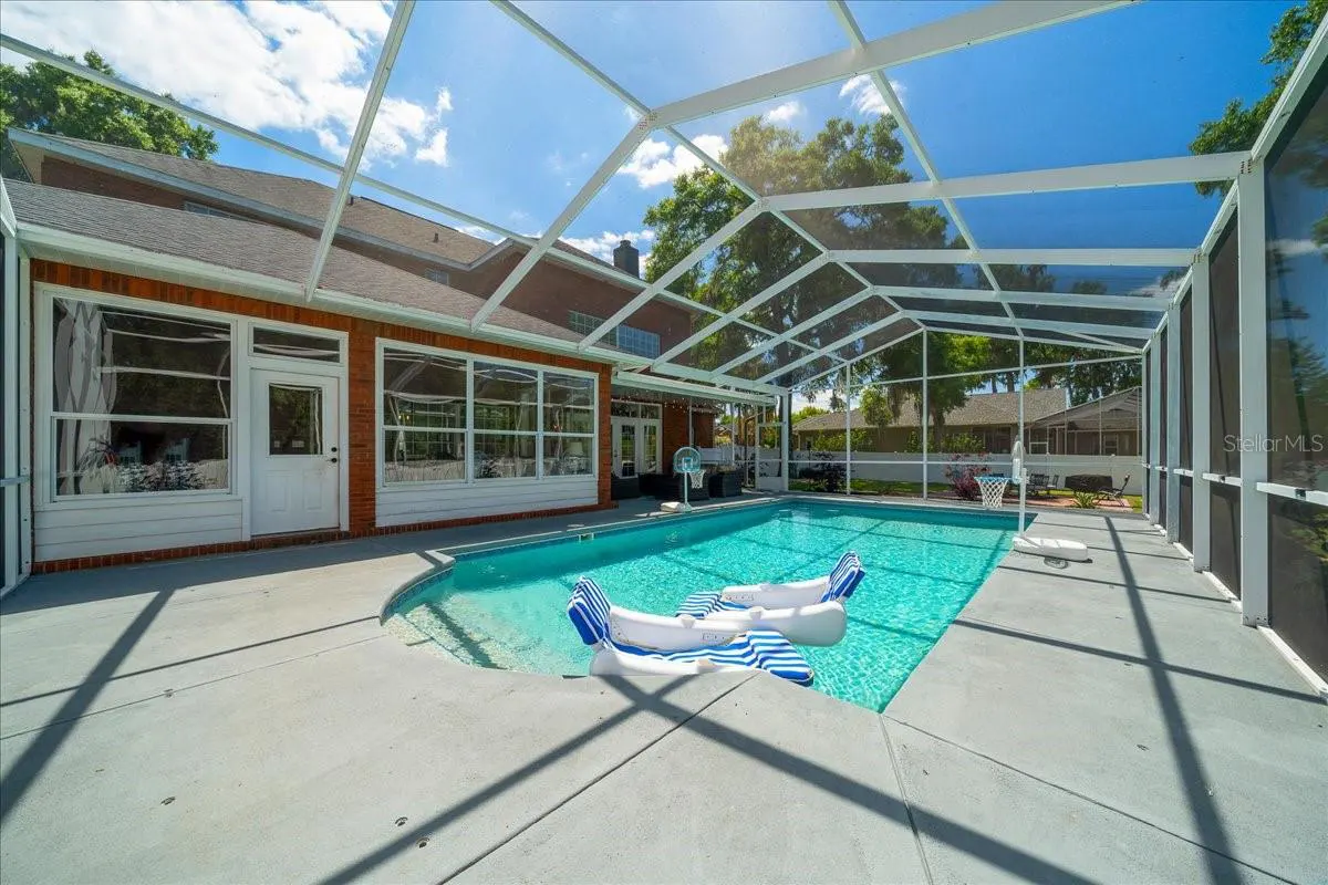 A screened-in pool with two airplane floats sits in a backyard with a brick house and blue sky.