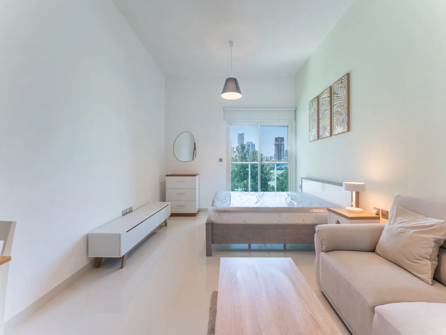 Bright studio apartment with white walls, beige sofa, bed, and city view through a large window. Modern, minimalist design.