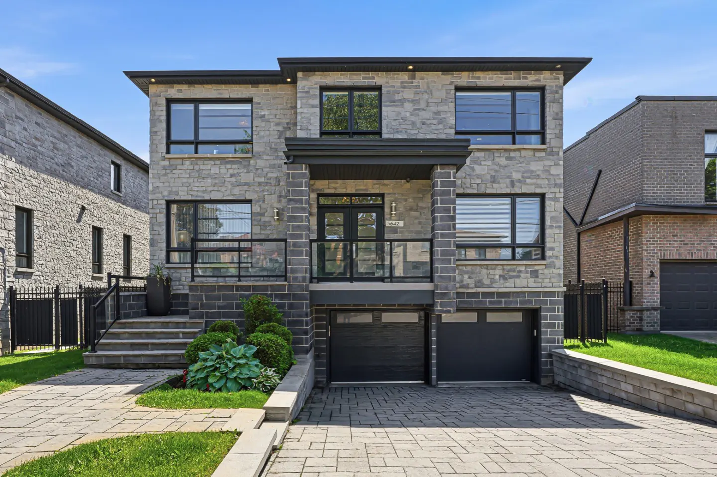 Modern two-story stone house with a double garage, glass-railed front porch, and paved driveway under a clear blue sky.