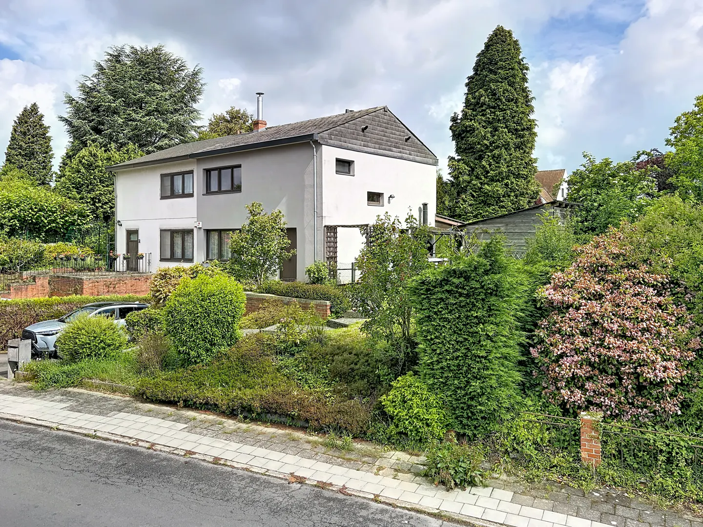 Two-story white house with dark trim, surrounded by lush greenery and trees under a cloudy sky. A car is parked in the driveway.