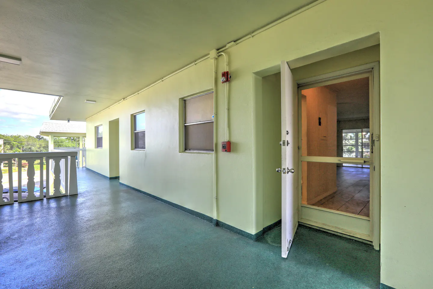 Exterior view of a light green apartment building hallway with an open white door and a blue-green floor. Windows and a balcony are visible.
