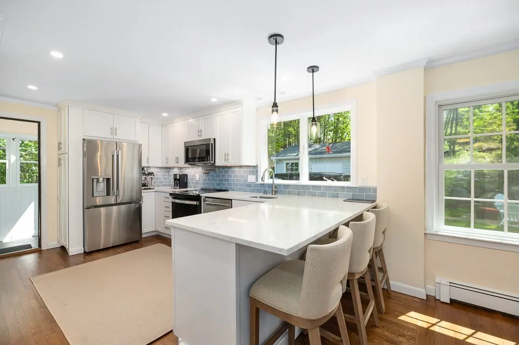 Bright kitchen with white cabinets, stainless steel appliances, and a white quartz island with three beige bar stools. A window overlooks a green backyard.