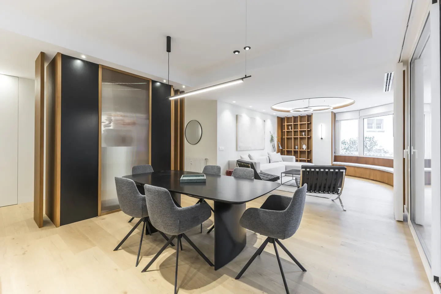 Modern open-plan dining and living area with a black table, grey chairs, light wood floors, and contemporary lighting.