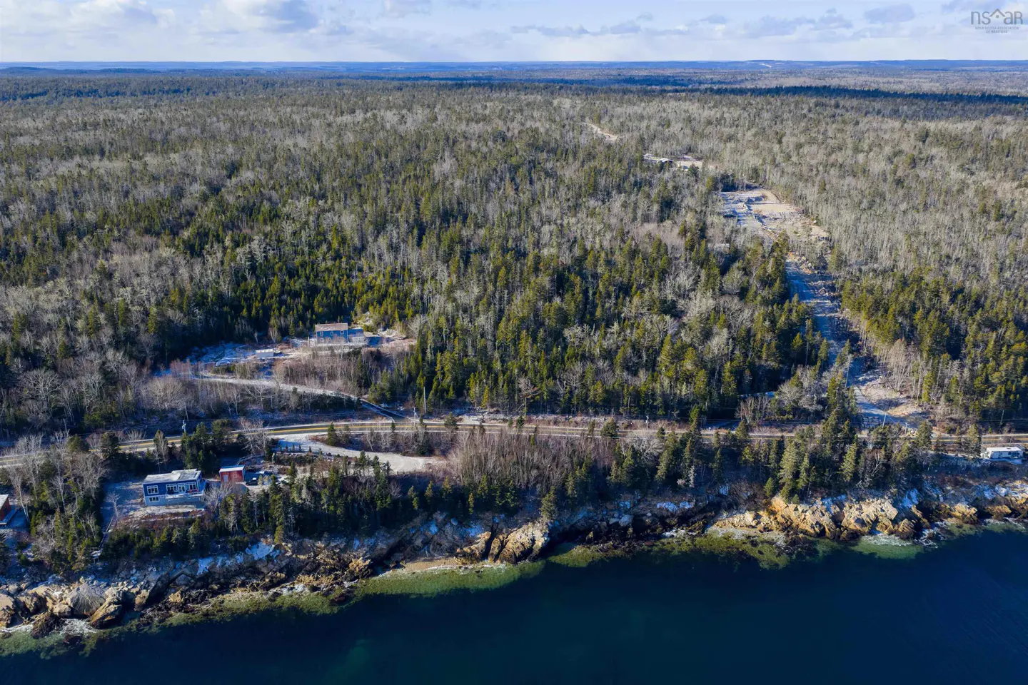 Aerial view of a coastal property with a house, dense forest, a road, and rocky shoreline meeting the deep blue ocean.