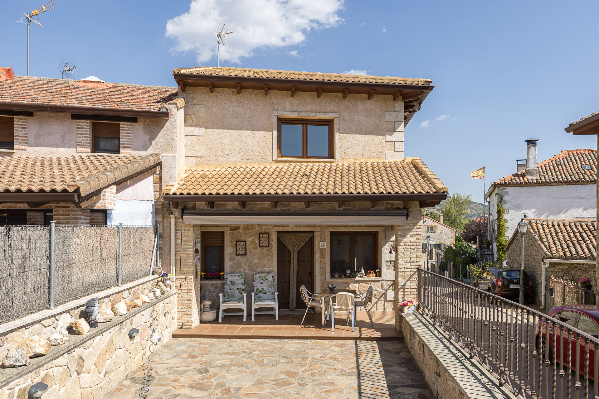 A two-story stone house with a tiled roof, featuring a front patio with chairs and a table under a covered porch. [AI-generated text]