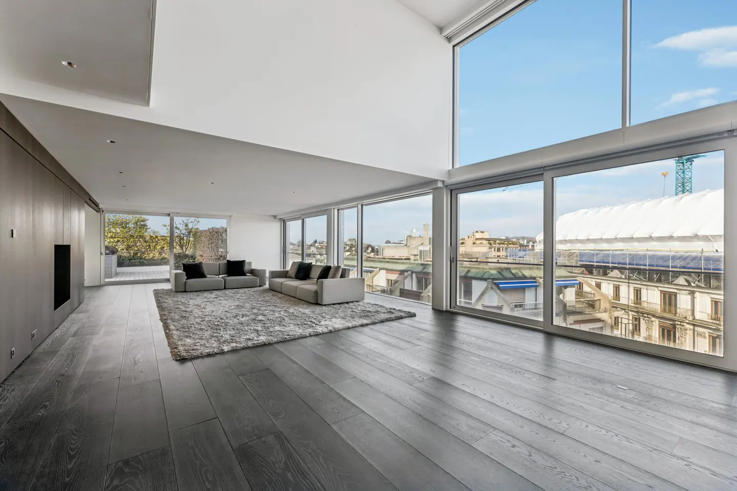 Bright, modern living room with dark wood floors, gray sofas, and large windows overlooking a city.