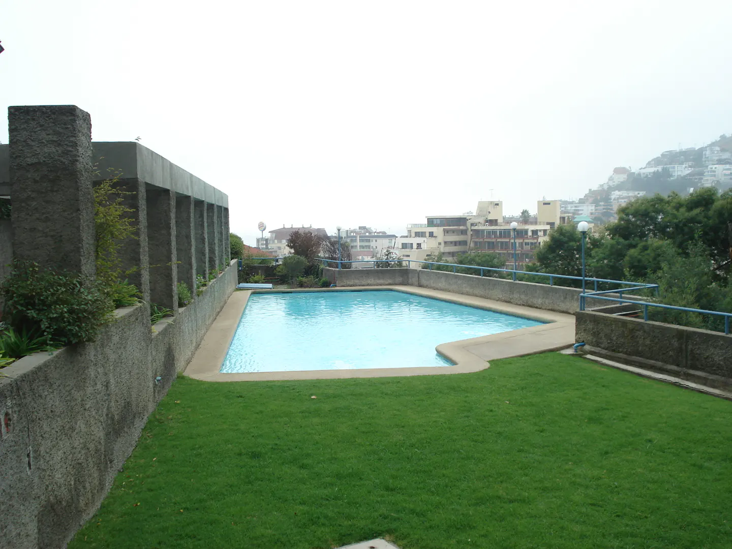 Outdoor pool with green lawn, concrete walls, and city views on a cloudy day.