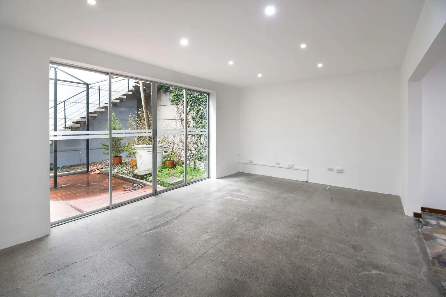 Bright, empty room with gray concrete floor and white walls. Large glass doors lead to a patio with plants and stairs.