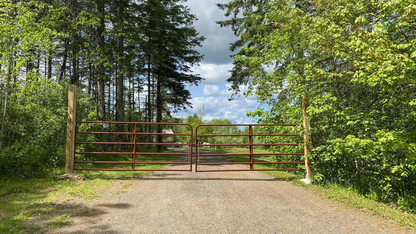 Gravel driveway leads to a red metal gate flanked by wooden posts and green trees. Blue sky with clouds visible beyond.