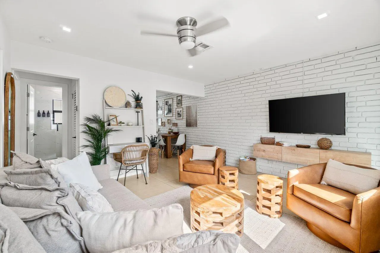 Bright living room with a white brick wall, TV, brown leather chairs, and a gray sofa with white pillows. A ceiling fan is visible.