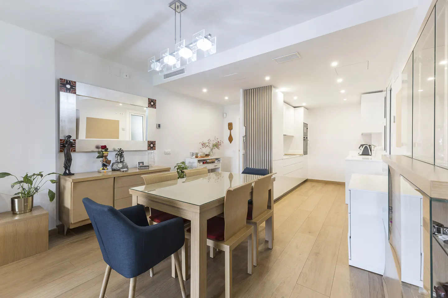 Modern dining area with a glass-top table, blue chairs, light wood floors, and an open-plan white kitchen in the background.