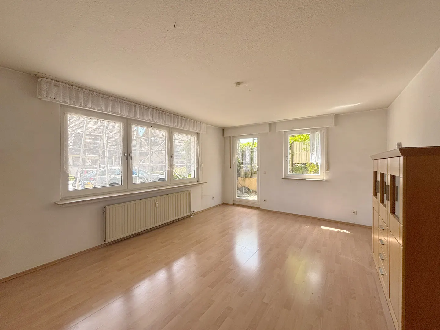 Bright, empty living room with light wood floors, white walls, large windows, a radiator, and a wooden cabinet.