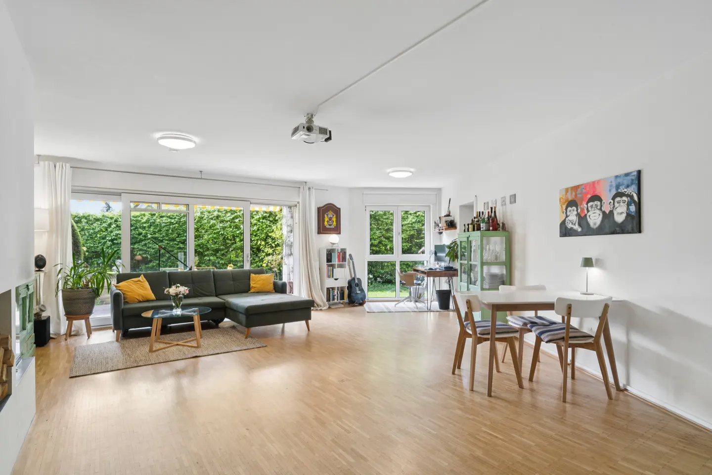 Bright, open living space with hardwood floors, a green sectional sofa, dining table, and a projector mounted on the ceiling.