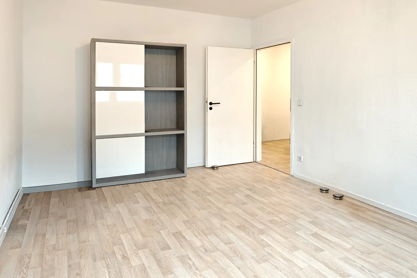 An empty room with light wood floors, white walls, and a gray and white bookshelf. A white door is ajar, leading to another room.