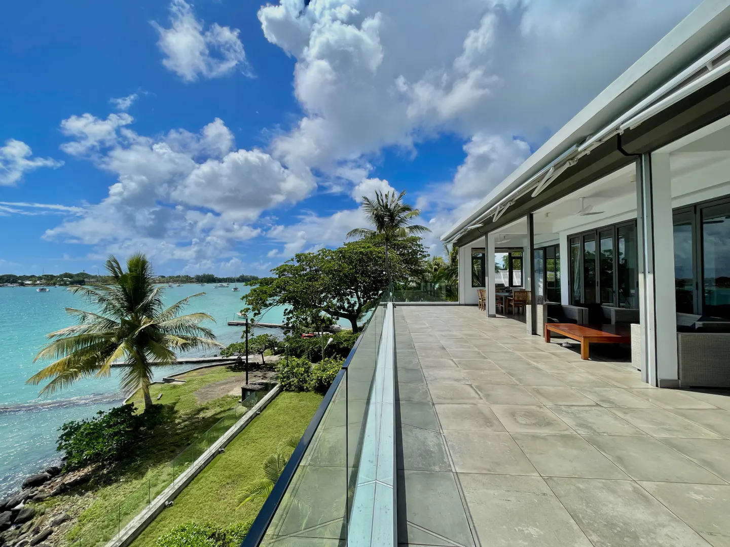 Waterfront property with a large patio, glass railing, and ocean view. Palm trees and green grass are visible. The sky is blue with white clouds.