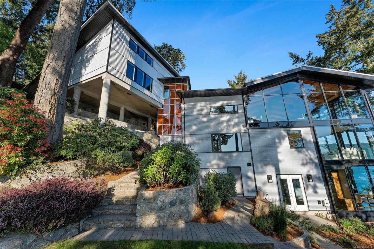 Exterior view of a modern, multi-level home with gray siding, large windows, and a stone pathway leading to the entrance. Lush greenery surrounds the house.