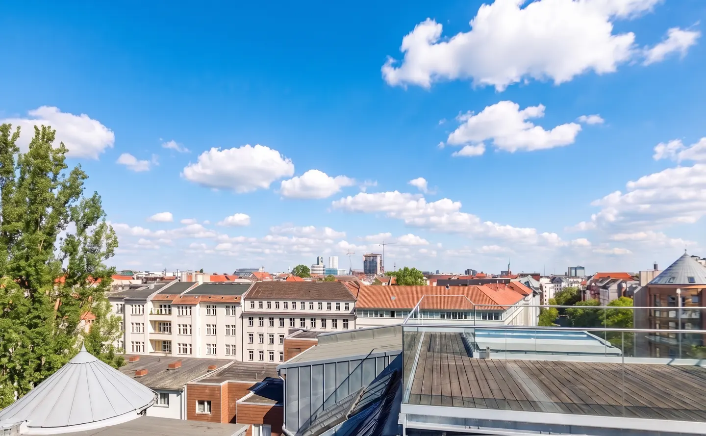 View from a rooftop with wooden planks, overlooking buildings with red roofs under a blue sky with white clouds.