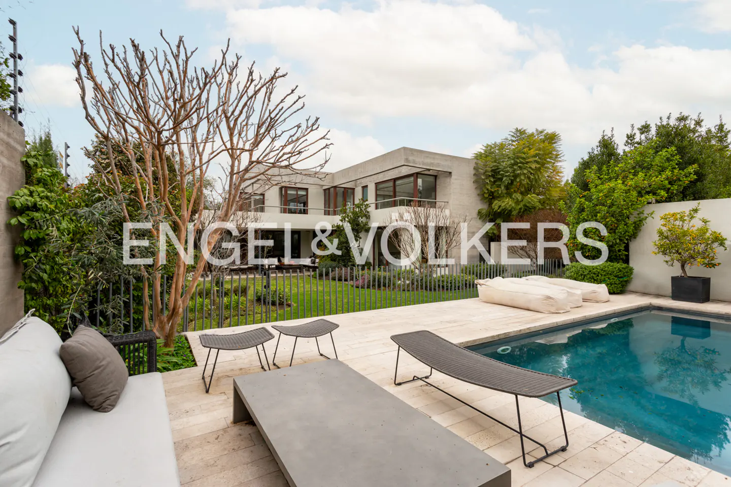 Backyard view of a modern house with a pool, lounge chairs, and a gray sofa. The sky is cloudy.