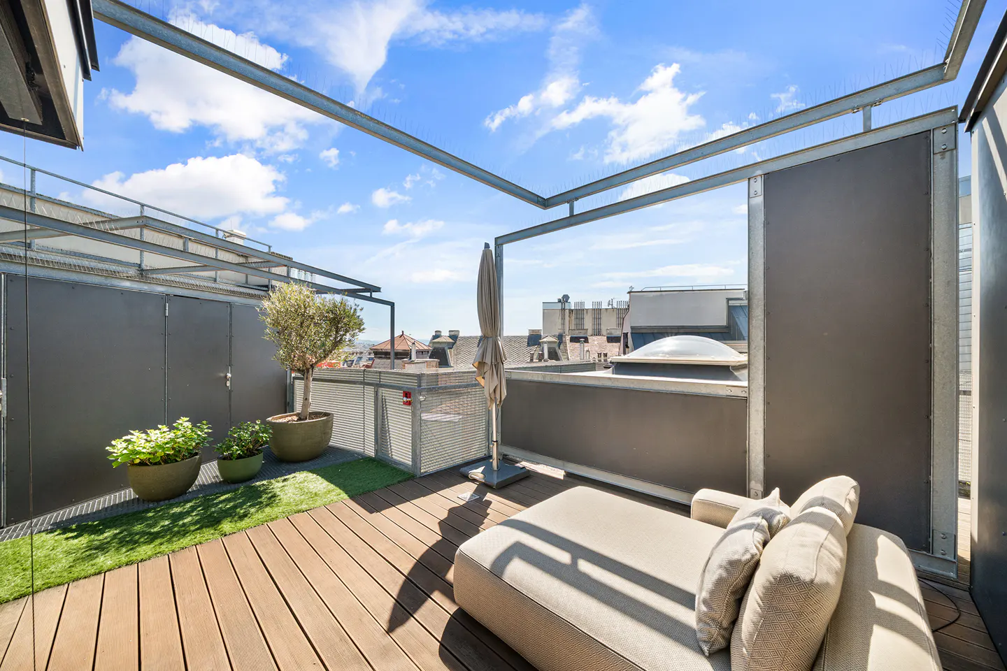 Modern rooftop terrace with wooden decking, a beige lounge chair, potted plants, and a metal frame under a blue sky.