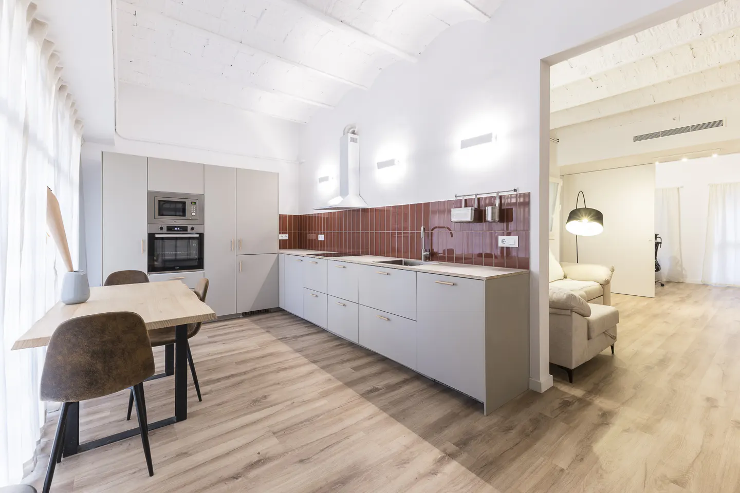 Bright, modern kitchen with light gray cabinets, red tile backsplash, and wood floors. A dining table and chairs are to the left. Living room visible.