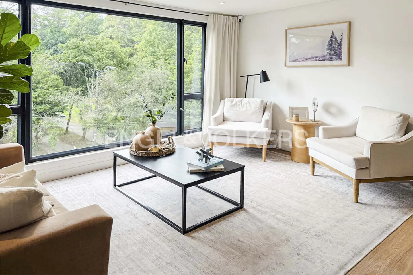 Living room with white walls, light carpet, and a black metal coffee table. Large window with green trees outside. Two white armchairs.