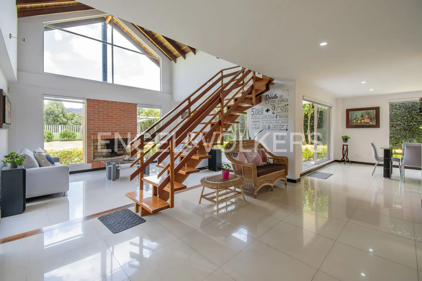 Bright, open-concept living room with wood staircase, brick fireplace, and wicker furniture on shiny tile floor. Large windows offer outdoor views.