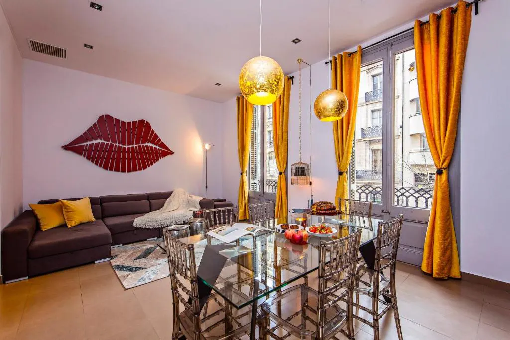 Living room with a glass table, clear chairs, and gold pendant lights. A brown couch with yellow pillows sits near a red lip art piece.