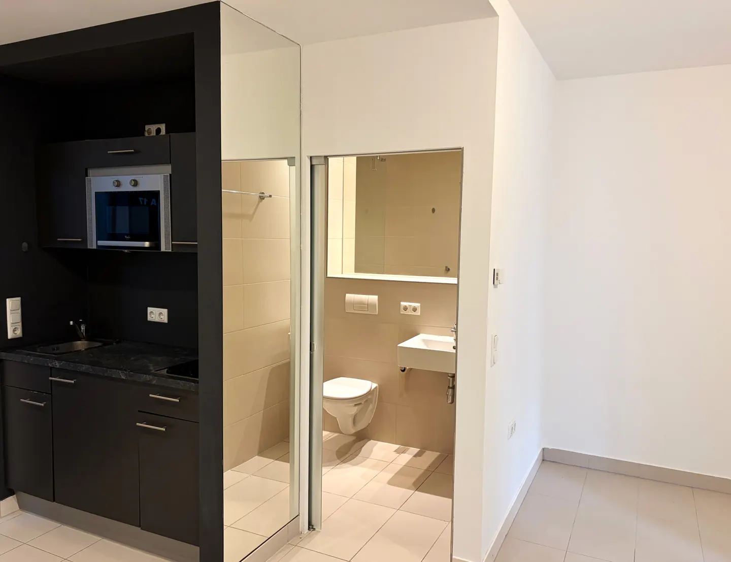 Studio apartment interior with black kitchenette, oven, and bathroom with toilet, sink, and shower. White walls and beige tile flooring.