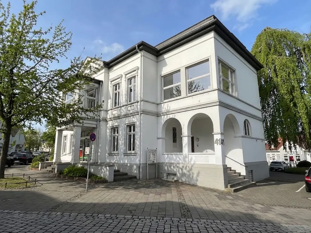 Two-story white building with arched entryway and gray trim, surrounded by trees and a brick-paved area.