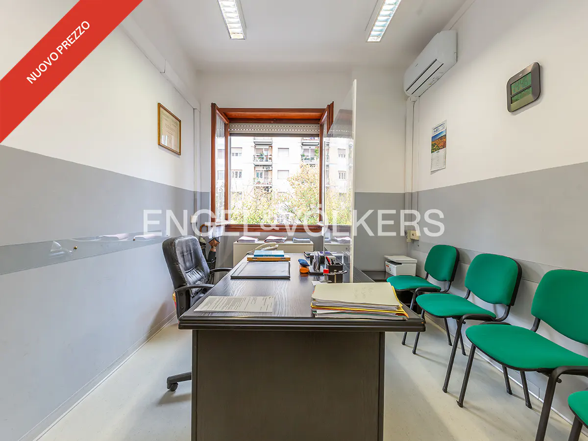 Office space with a dark desk, black chair, and green guest chairs. A window overlooks a city street. "Nuovo Prezzo" banner in the corner.