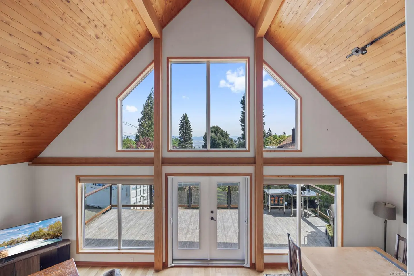 Bright living room with wood ceiling, white walls, and large windows. Outside, trees and a blue sky are visible. A deck is accessible through glass doors.