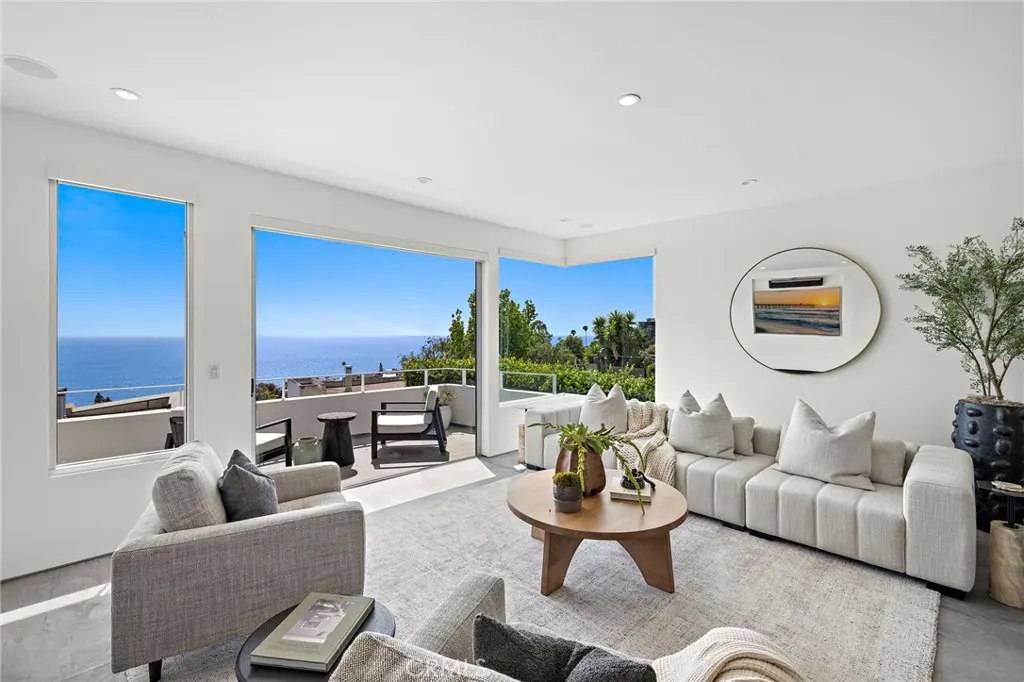 Modern living room with white walls, neutral furniture, and a large balcony overlooking the ocean and blue sky.