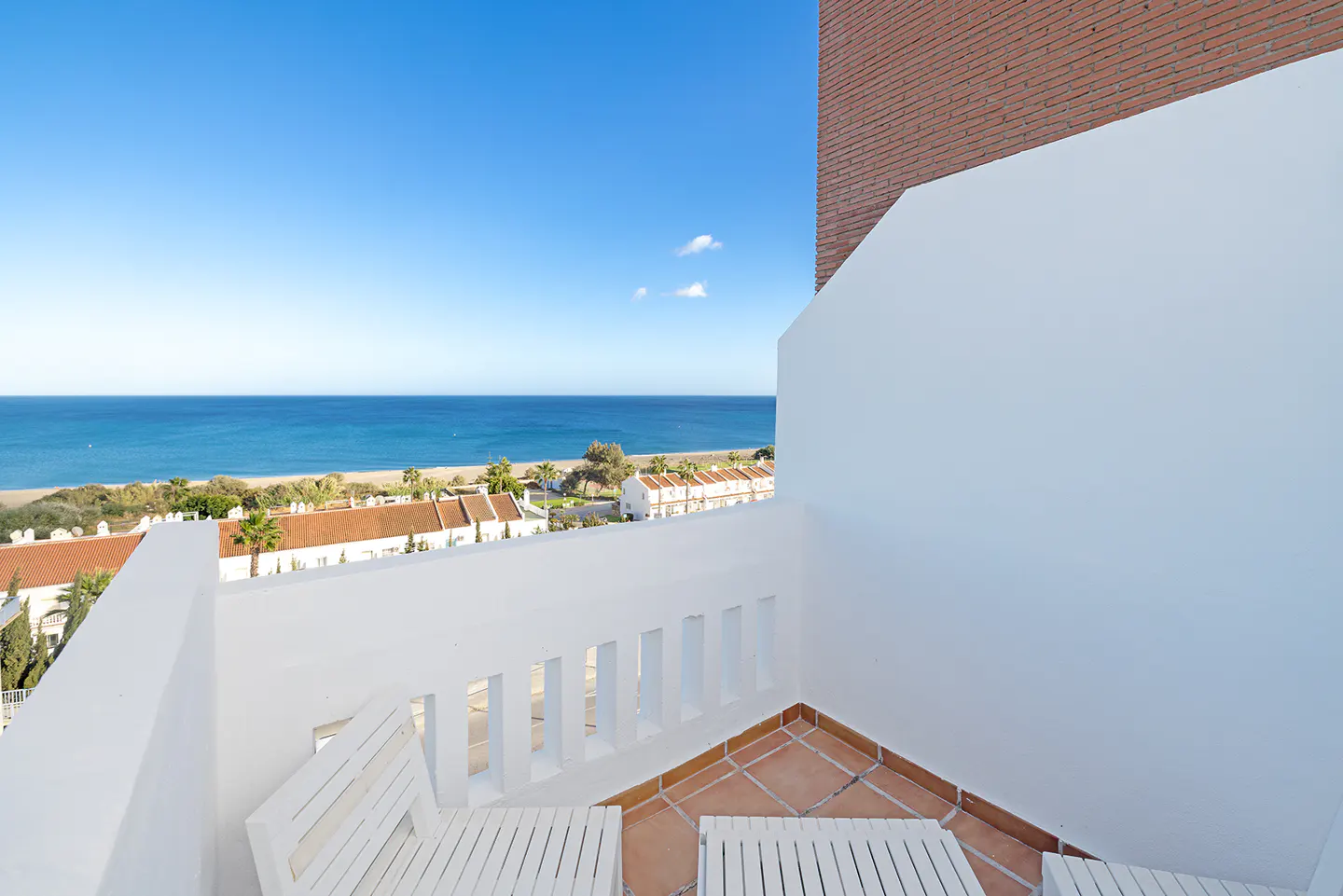 View from a white balcony with chairs overlooking the ocean, beach, and houses with orange roofs. Blue sky.