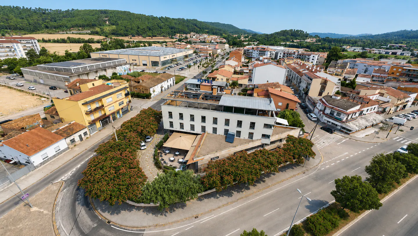 Aerial view of a white hotel building surrounded by trees and roads in a small town with hills in the background.