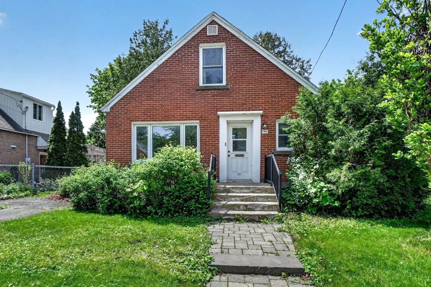 A one-story red brick house with a white front door, stone steps, and a stone walkway surrounded by green bushes and lawn.