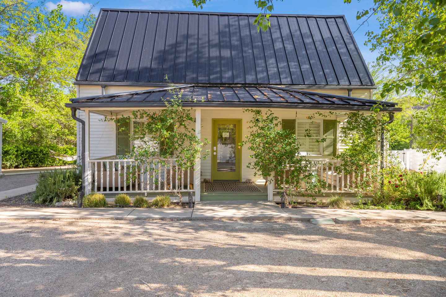 Exterior view of a white house with a black metal roof, green door, and a small porch with climbing plants.
