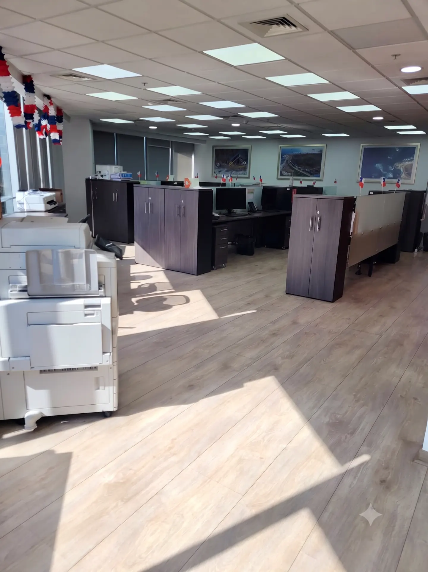 Spacious office with light wood floors, dark storage cabinets, cubicle desks, and a large white printer in the foreground.