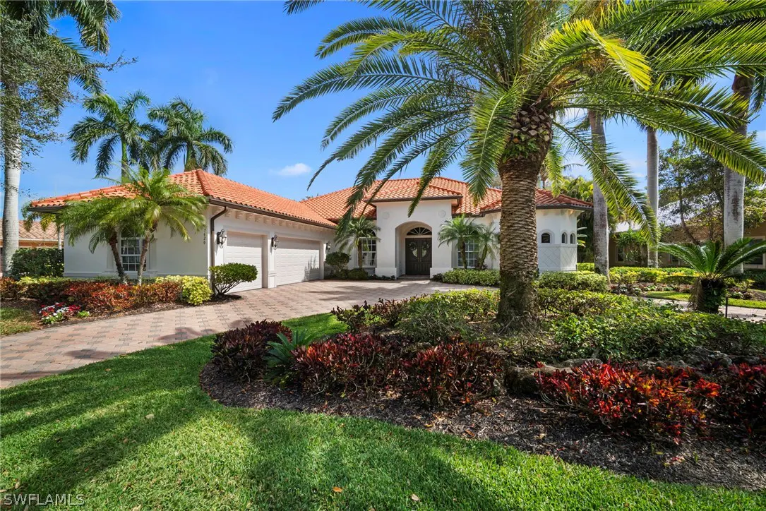 A white house with a red tile roof, palm trees, and a brick driveway on a sunny day.