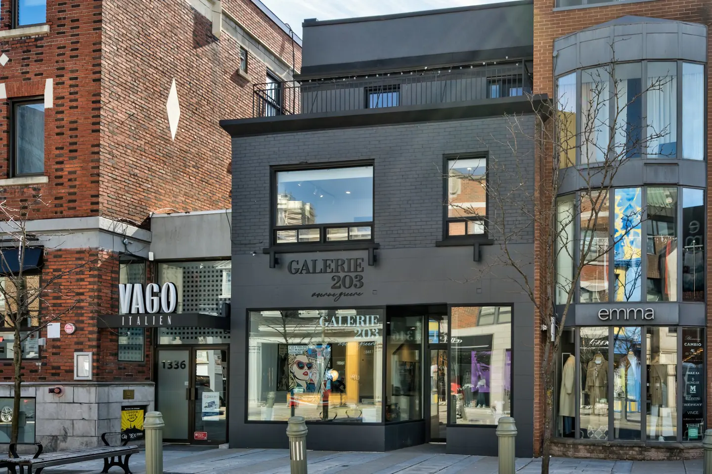 Street view of Galerie 203, a two-story black brick building, nestled between Vago Italien and Emma clothing stores.