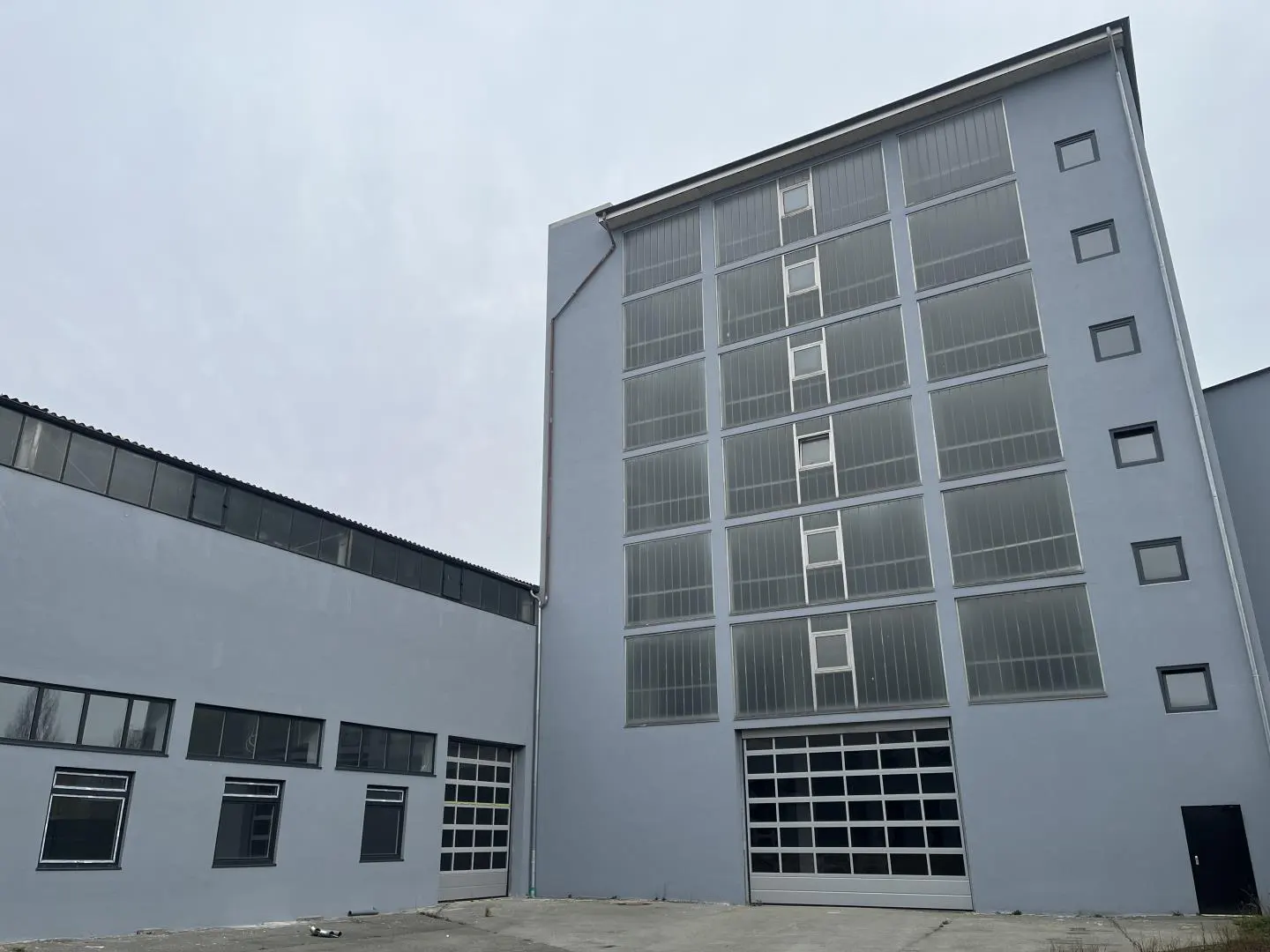 Exterior of a gray industrial building with large garage doors and many windows under a cloudy sky.