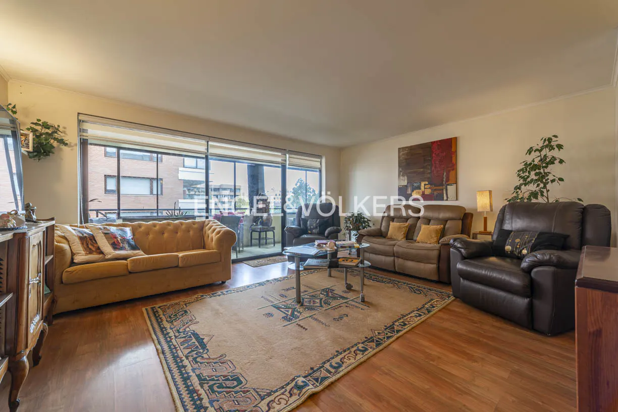 Living room with hardwood floors, a patterned rug, and tan sofas. A balcony is visible through a large window.