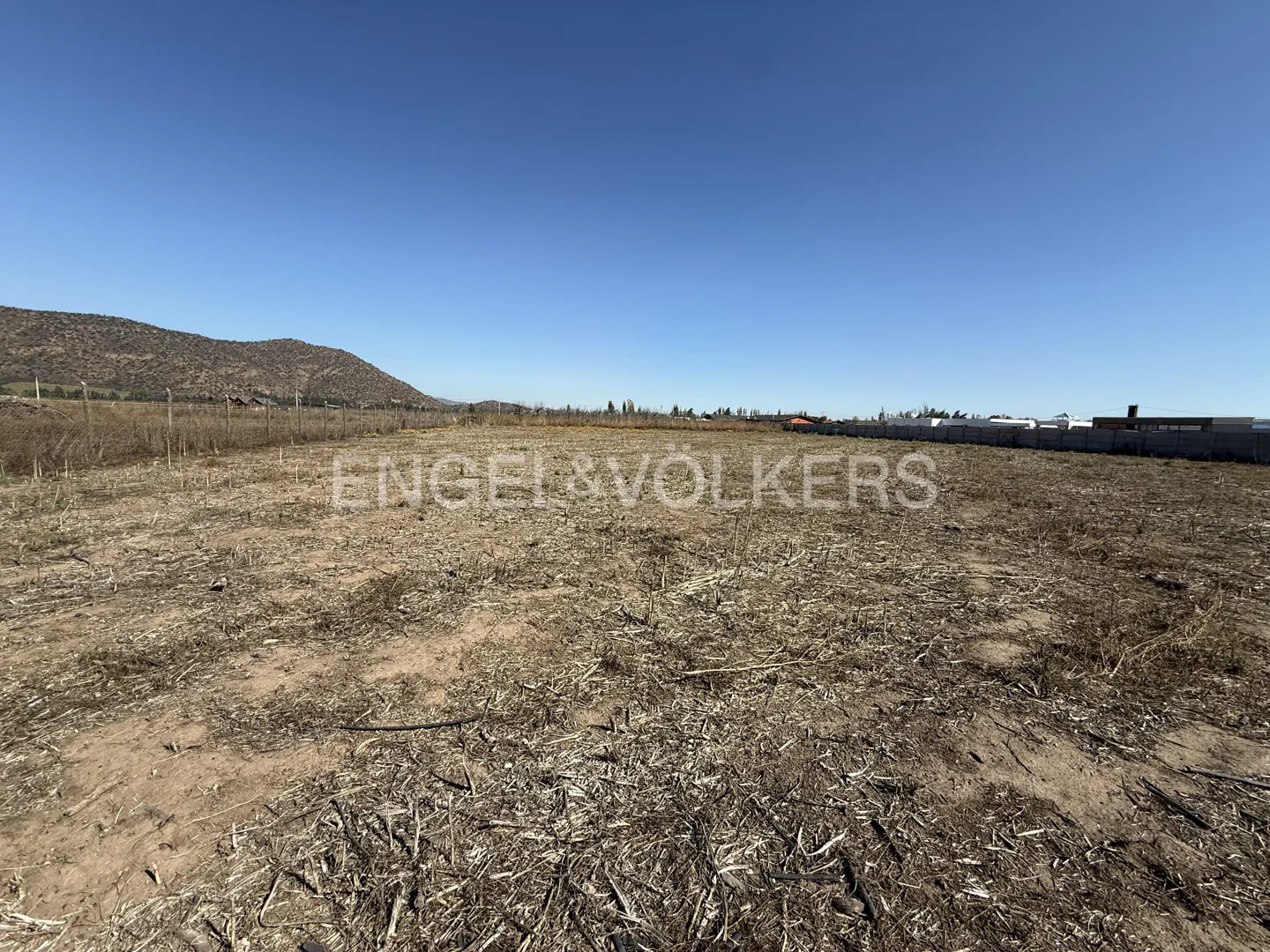 Vacant lot with dry grass under a clear blue sky. Distant hills and a fence line the property. Engel & Völkers logo overlay.