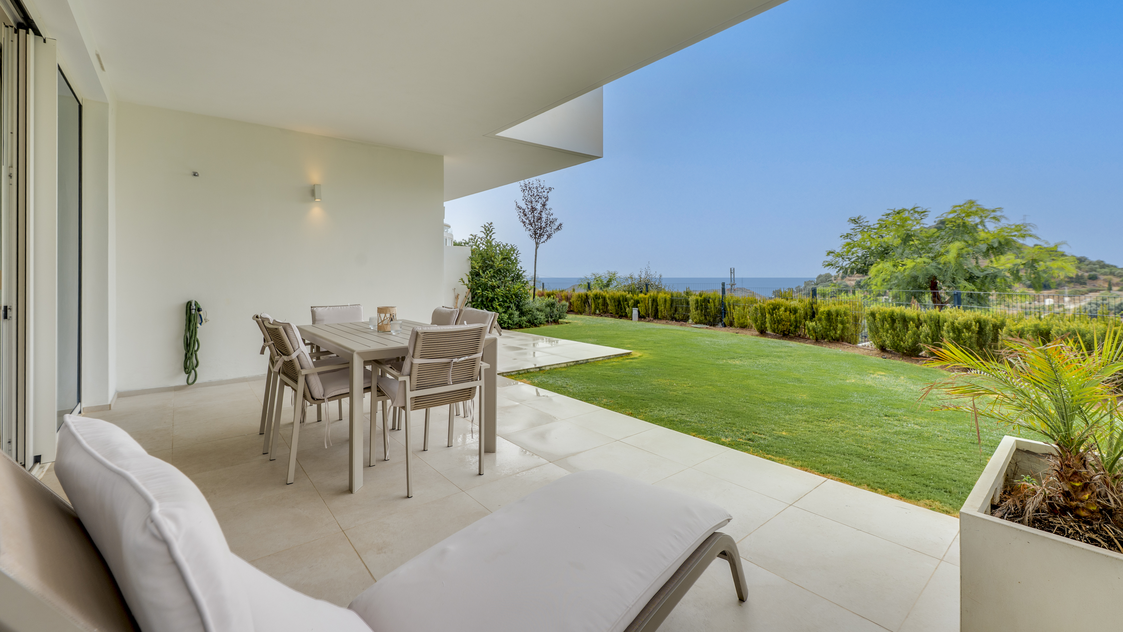 Modern covered patio with dining set and lounge chair overlooking a green lawn, manicured hedges, and distant sea view.