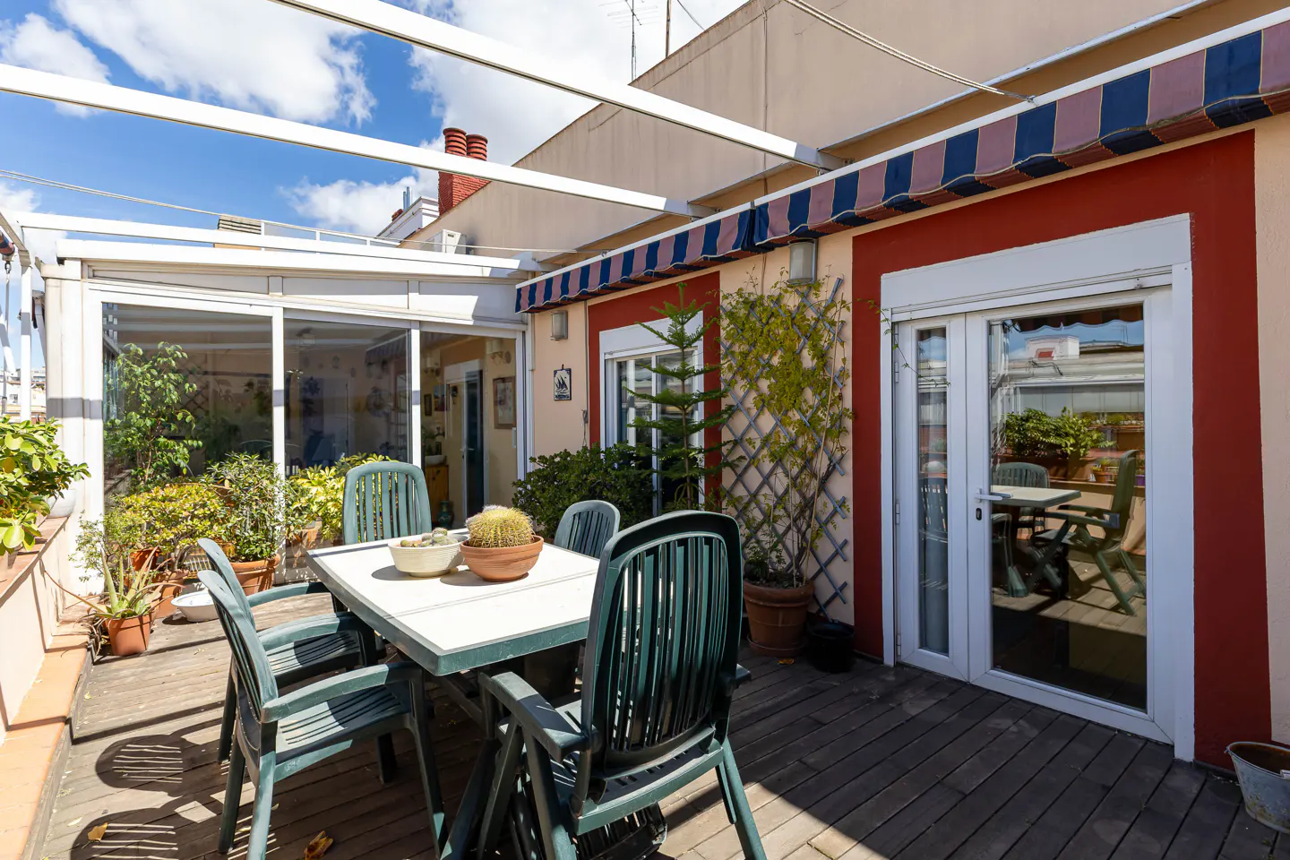 Outdoor patio with a white table, green chairs, and potted plants on a wooden deck. Awnings and a glass-enclosed room are in the background.