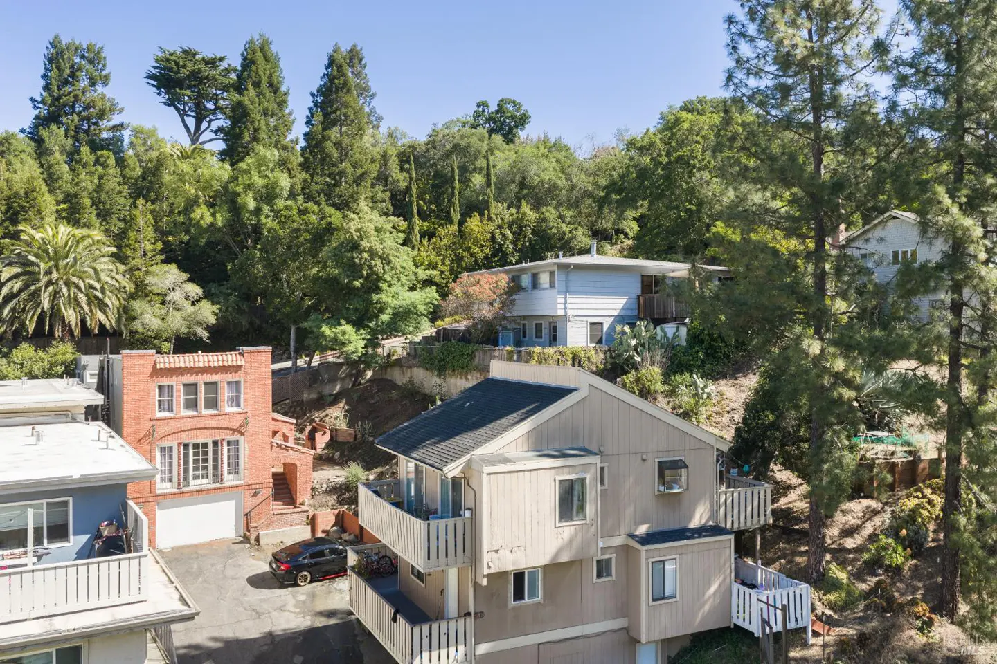 Aerial view of multi-level homes on a wooded hillside, featuring a red brick house and a beige house with balconies.