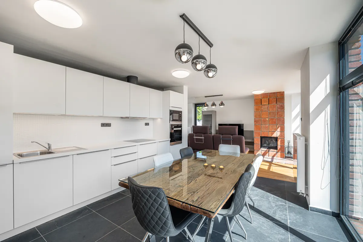 Bright, modern kitchen and dining area with white cabinets, wood table, and black chairs. A brick fireplace is visible in the background.
